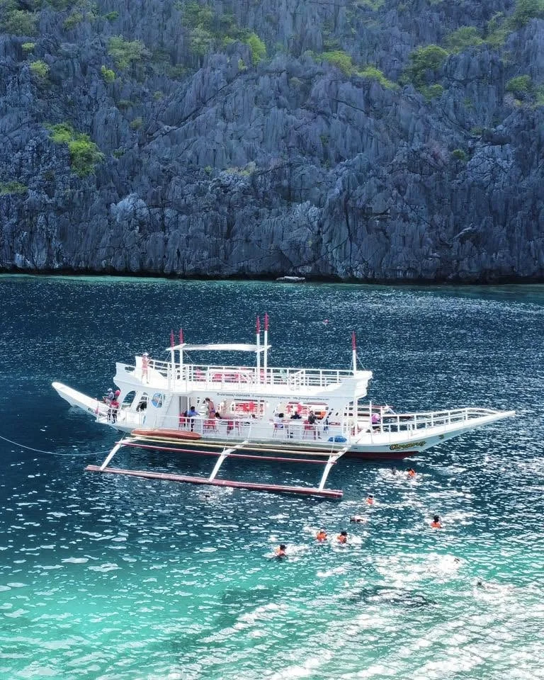 The two-deck Anang premium bangka anchored in turquoise water below limestone cliffs, with guests snorkeling nearby
