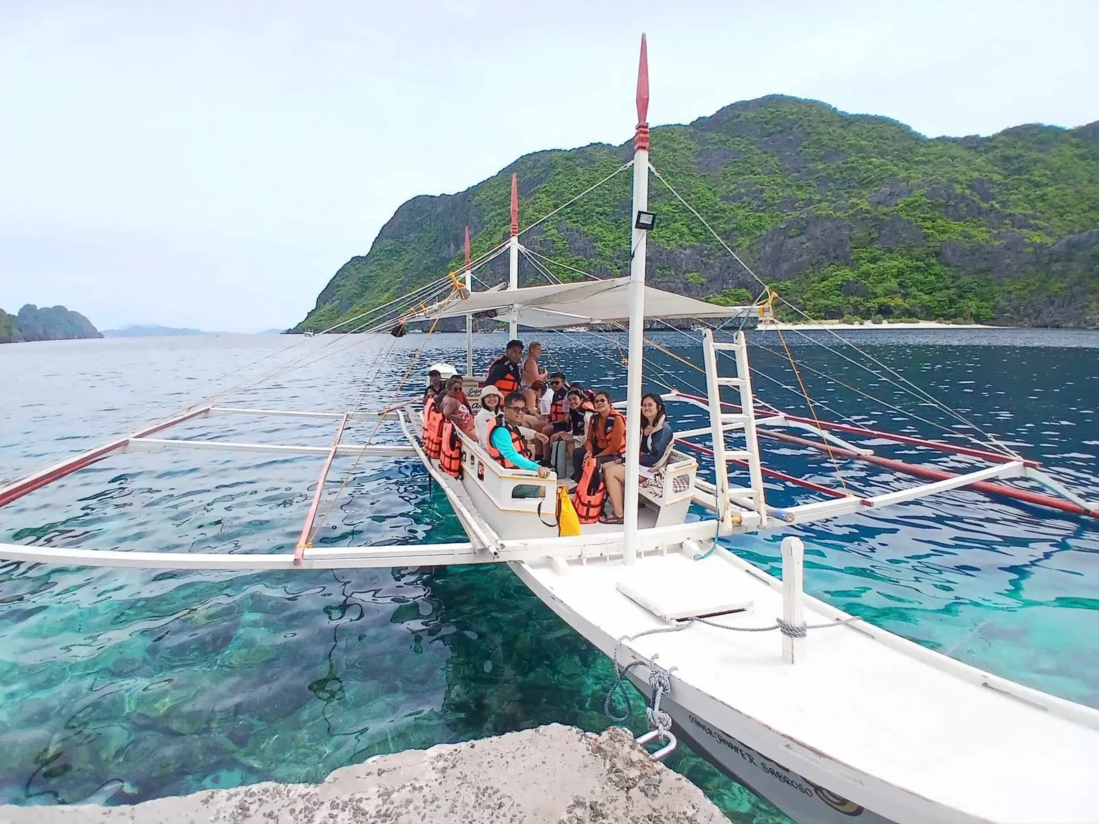 Guests in life vests aboard a traditional Anang bangka beside a pier, with clear water and limestone islands